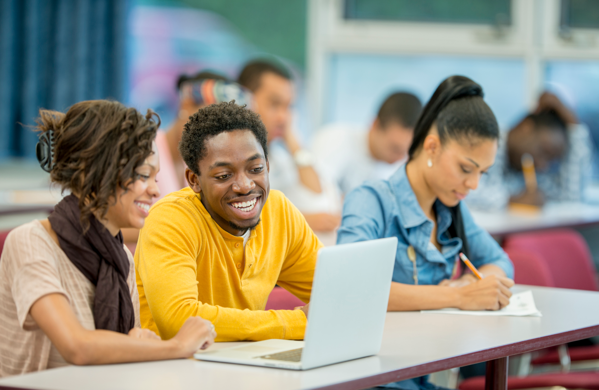 Three students sitting at a desk, with two students smiling and sharing a laptop.